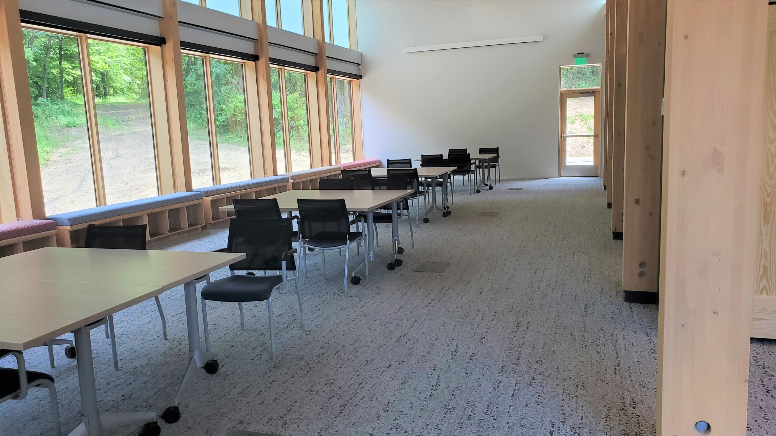 Multipurpose room with large windows, wood beams, light wooden tables, and black chairs