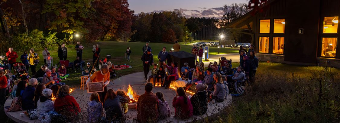 A fall Parks evening program with people sitting around a bonfire pit listening to presenter.